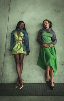 Two stylish women posing in trendy, modern green dresses against a concrete backdrop.