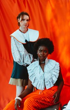 Fashion-forward portrait of two women in unique, colorful outfits against an orange backdrop.