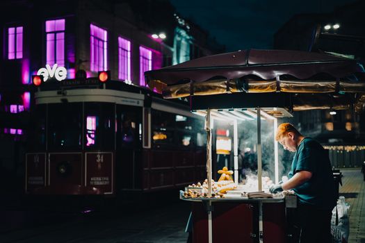A street vendor preparing food in bustling nighttime Istanbul with tram and neon lights backdrop.