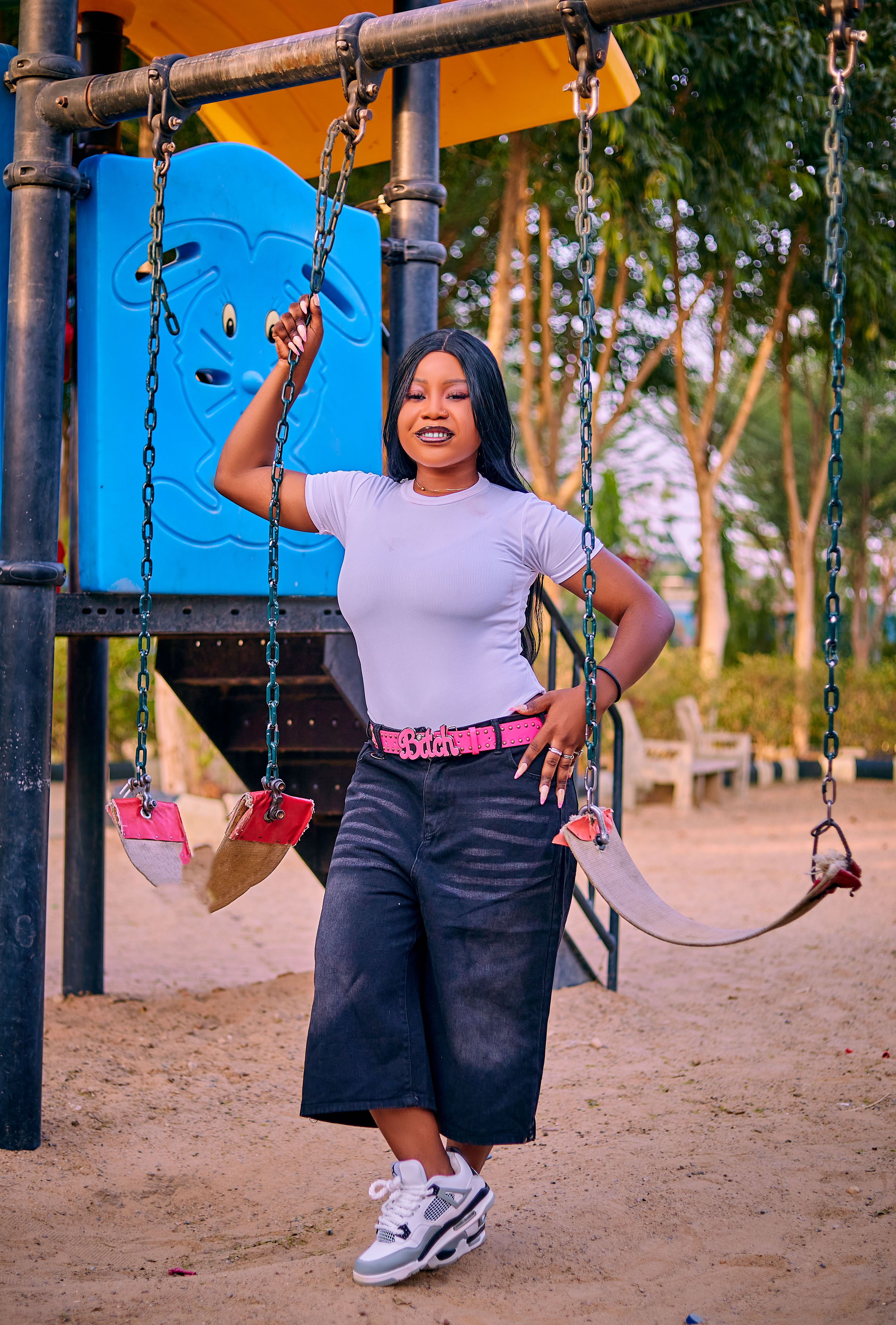 A stylish woman poses confidently next to a swing set in a playful outdoor park setting.