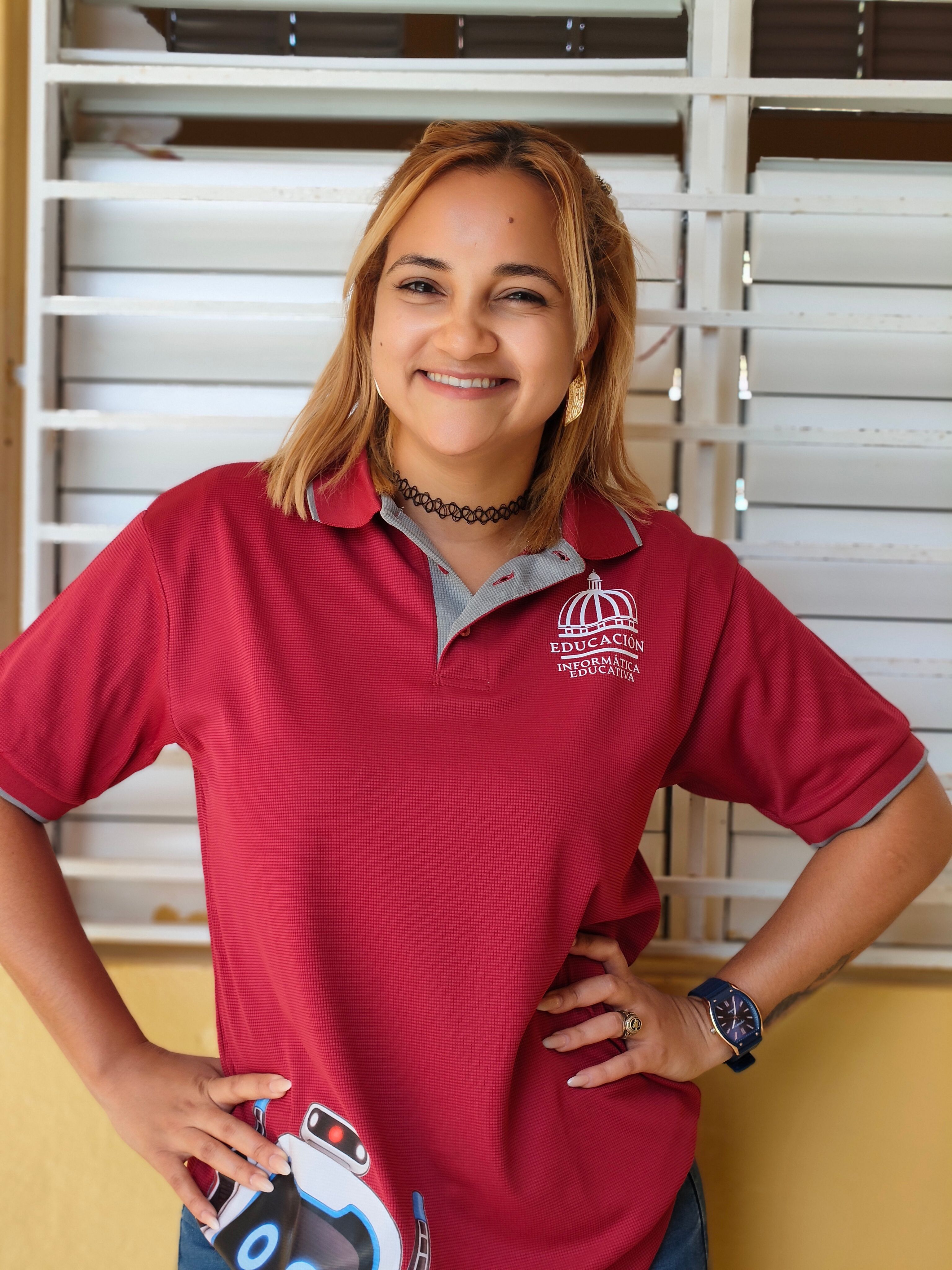 A cheerful woman stands confidently in a red polo shirt, indoors.