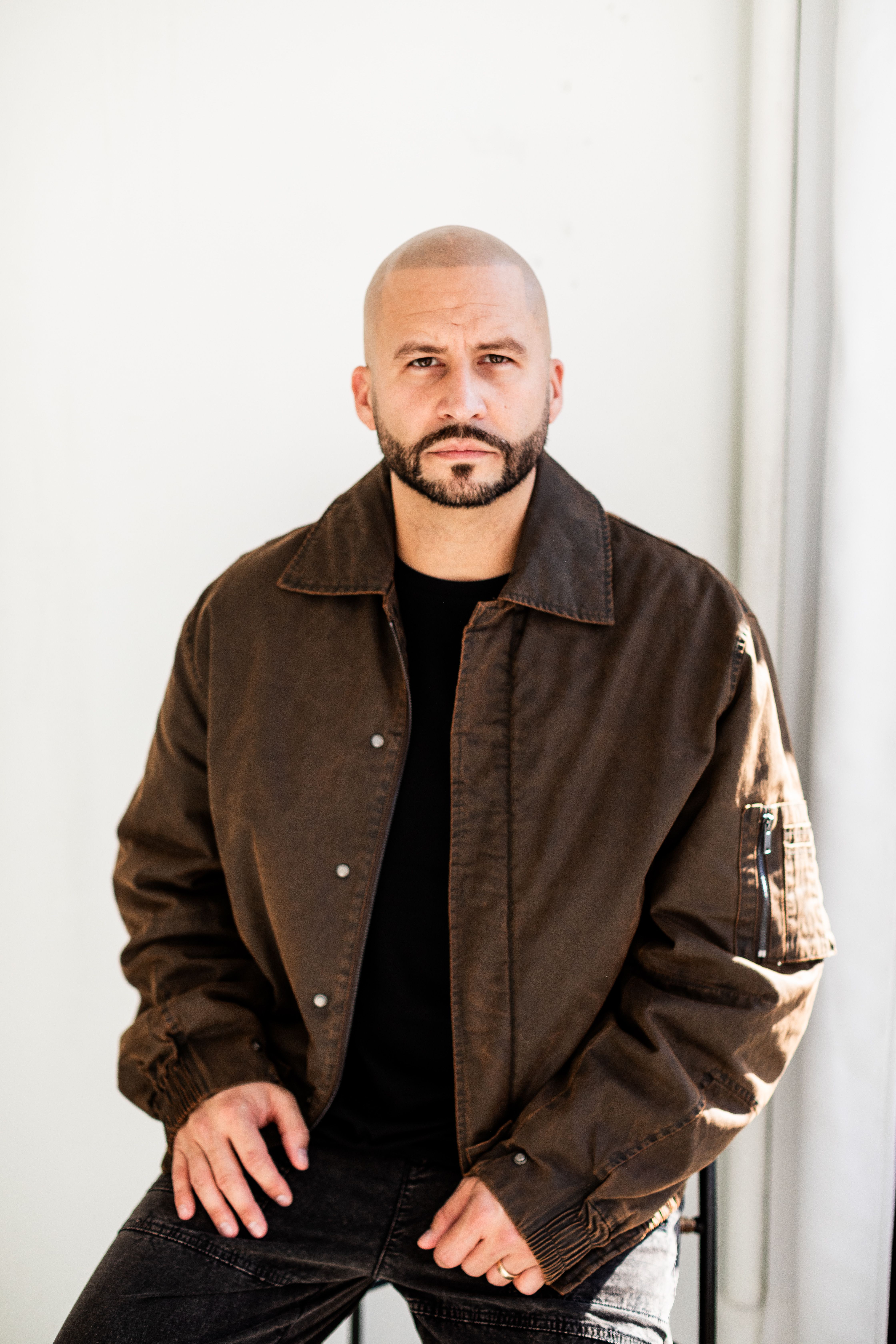 A confident man in a brown jacket poses indoors with soft natural lighting.