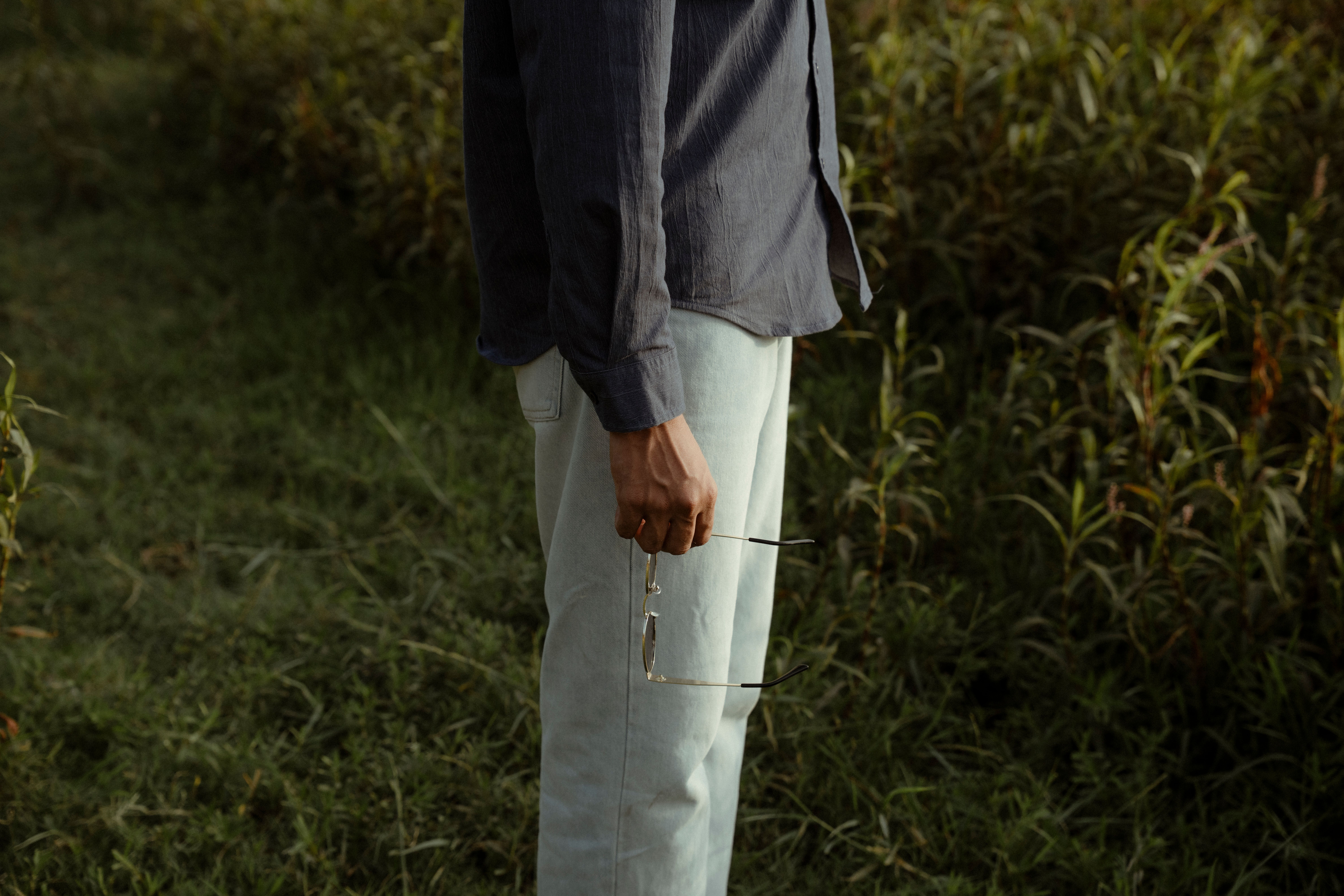 A person in casual attire holding sunglasses outdoors in a grassy field.
