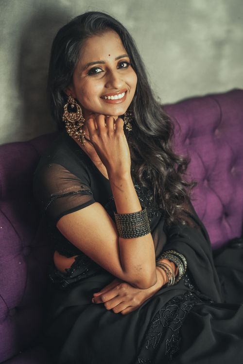 Free Beautiful woman in black saree seated on purple couch with warm smile. Stock Photo