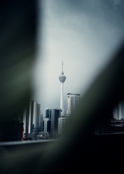 Silhouetted Kuala Lumpur Tower amidst city skyscrapers viewed between blurred foreground elements.