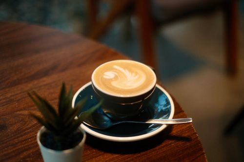 Free A warm cup of latte with artistic foam sits on a table in a cozy cafe, exuding warmth. Stock Photo