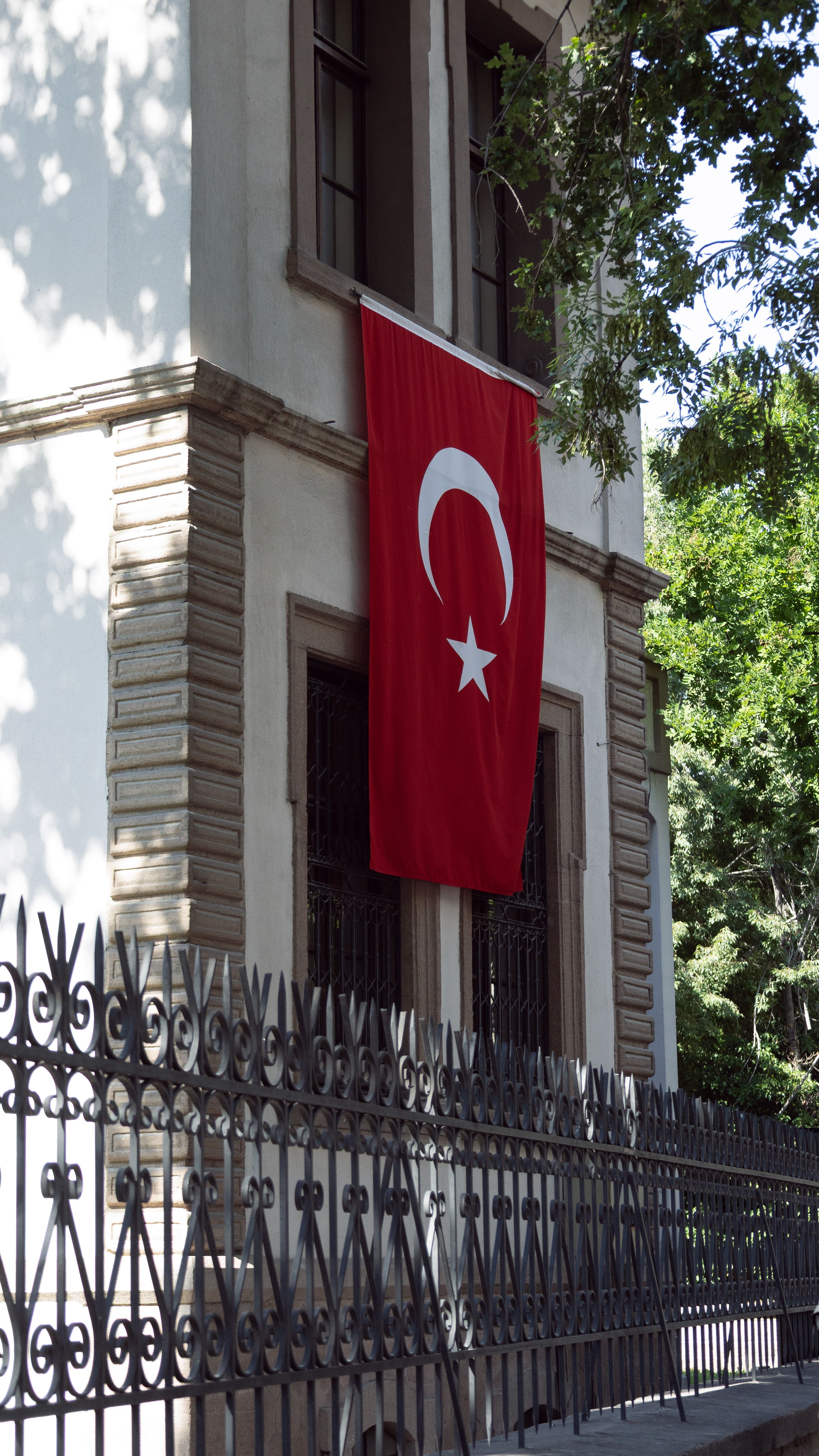 A Turkish flag displayed on a historic building facade in Konya, Türkiye, under sunlight and surrounded by trees.