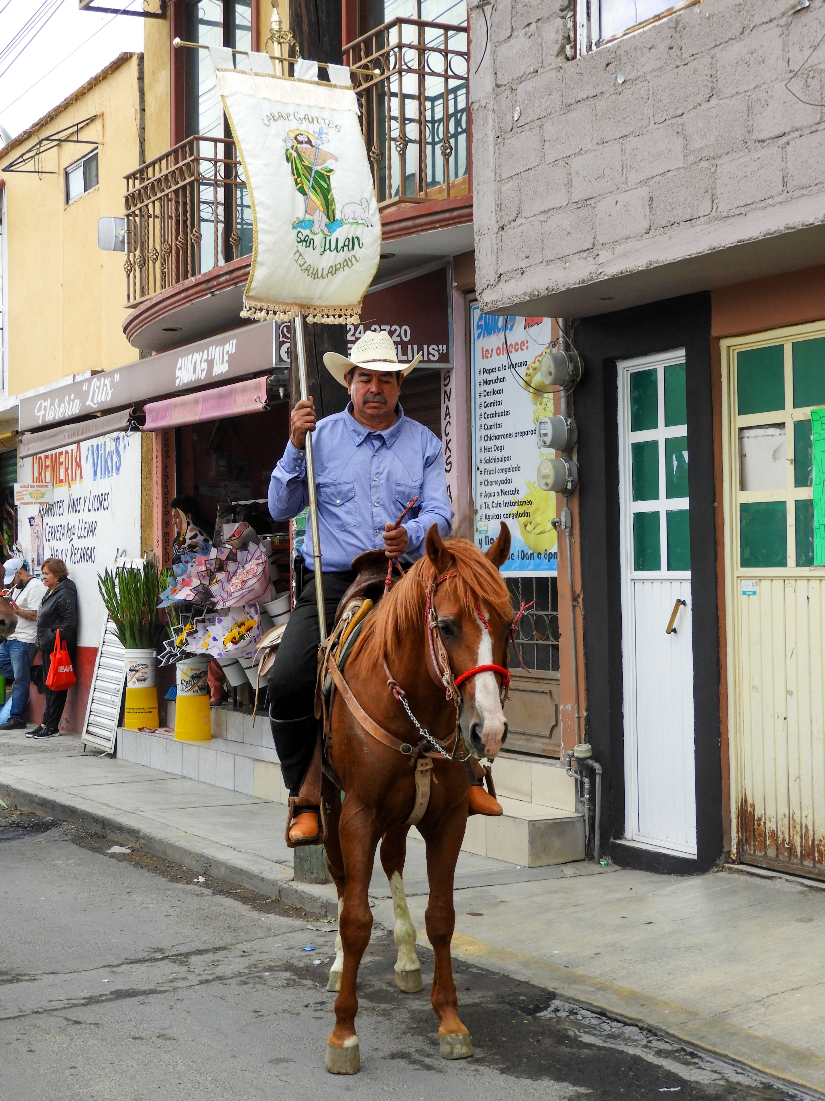 A man on horseback holding a banner during a Mexican street parade, showcasing traditional attire.