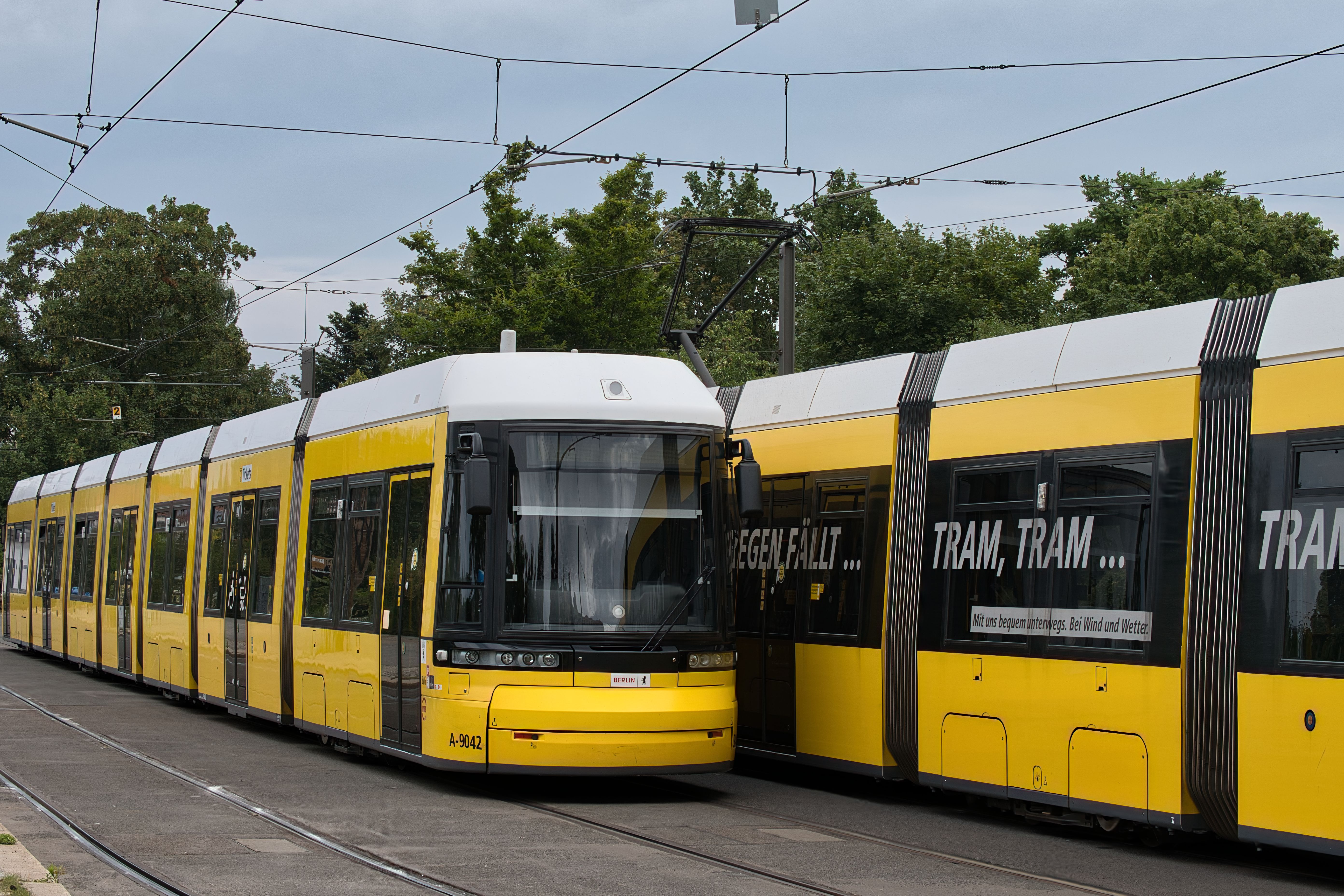 A yellow tram moves through a city street near greenery under cloudy skies.
