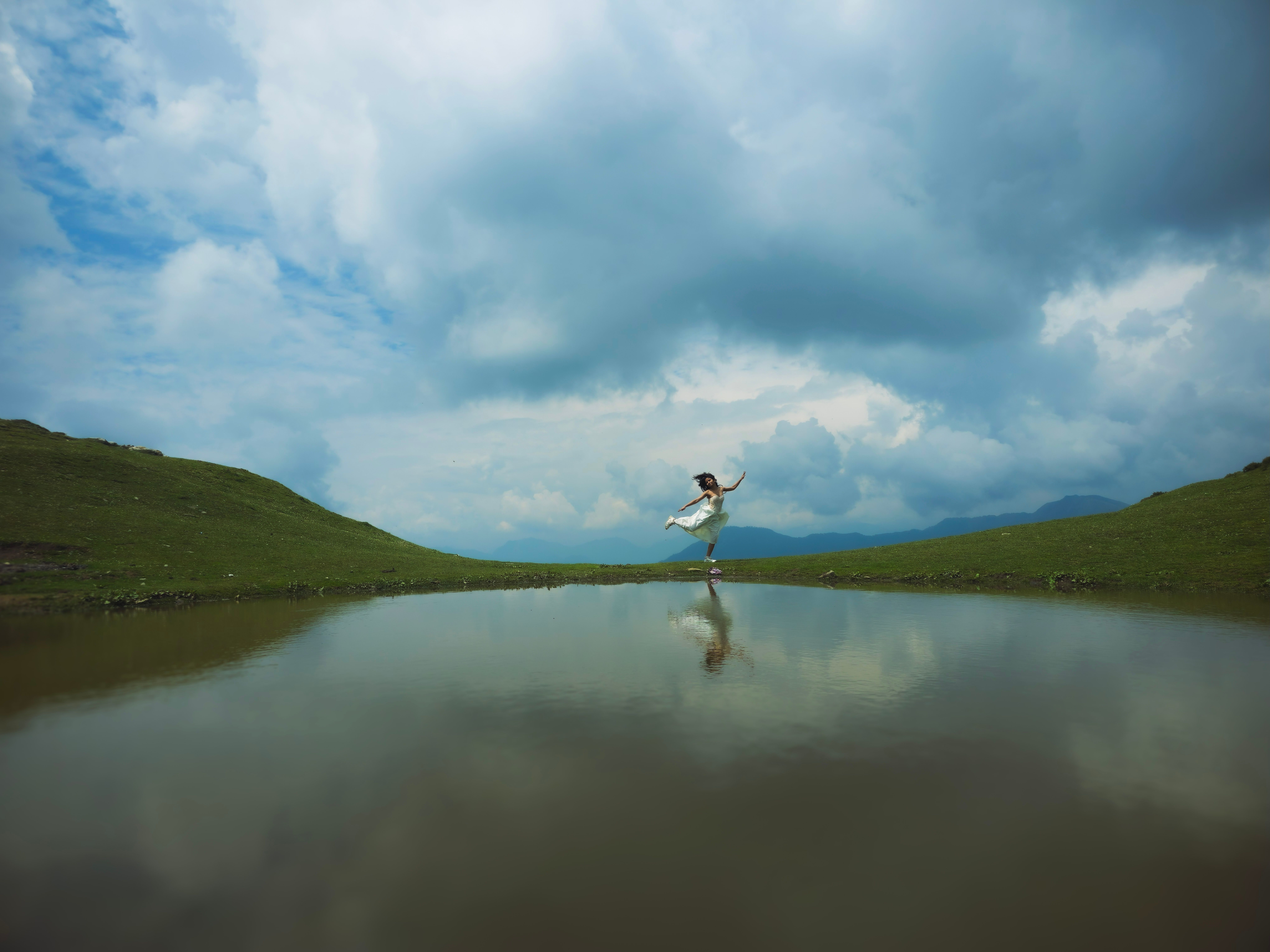 Serene landscape with a woman in white dress reflecting on calm lake surrounded by clouds.
