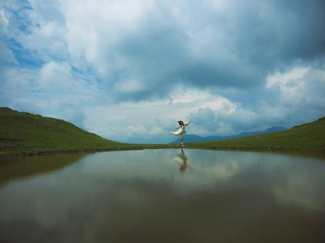 Serene landscape with a woman in white dress reflecting on calm lake surrounded by clouds.