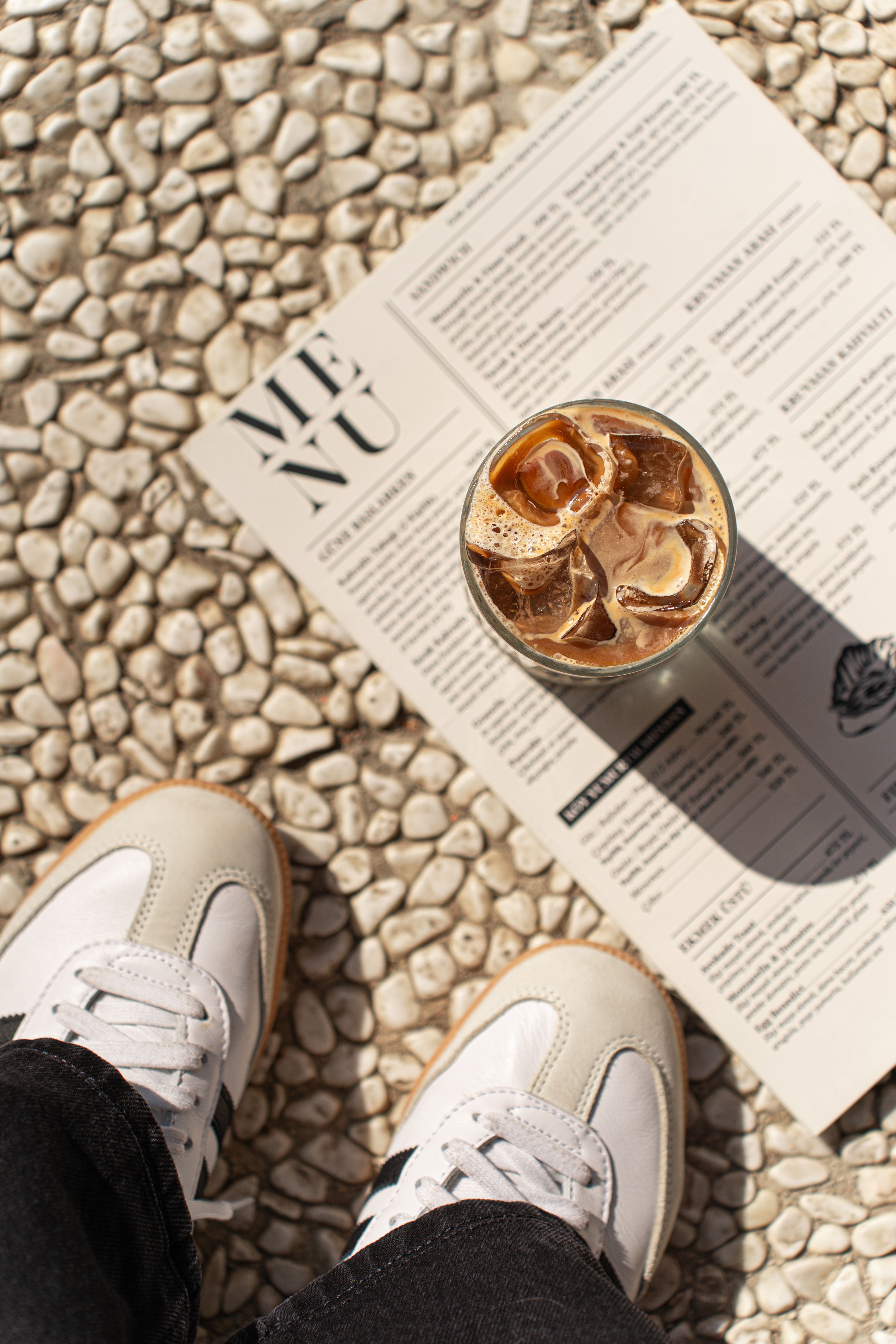 Flat lay of iced coffee on menu with sneakers on pebbled ground, perfect for coffee culture themes.