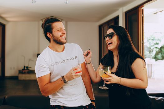 Delighted young man and woman in casual clothes laughing and talking with each other while drinking cocktails in modern apartment on weekend
