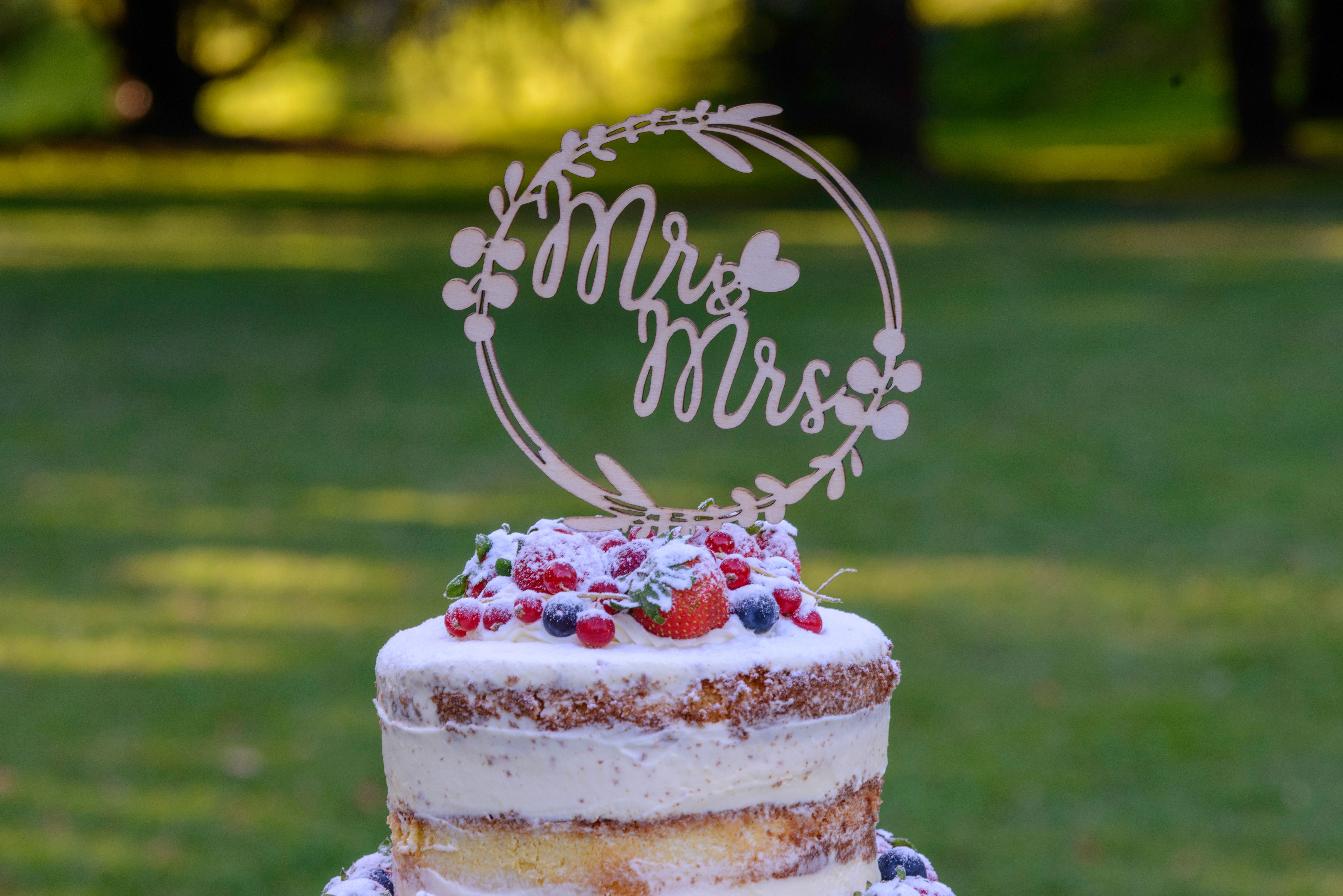 A rustic naked wedding cake adorned with fresh berries and a 'Mr & Mrs' topper in an outdoor setting.