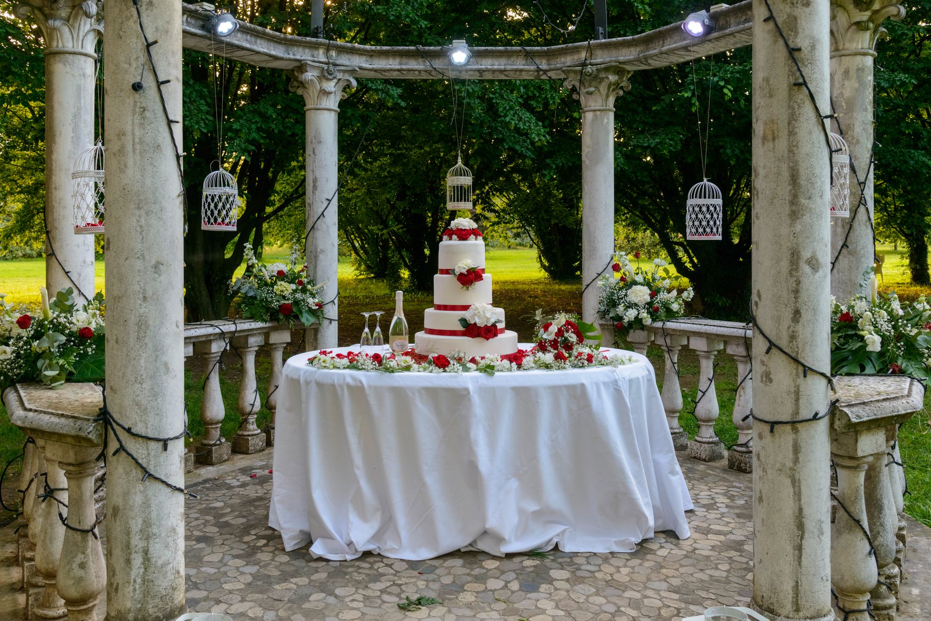 Kue pengantin bertingkat elegan dengan mawar merah di lokasi luar ruangan yang indah.