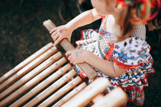 From above of cute kid in vivid colorful dress sitting on ground and holding to arm of wooden chair during holidays in garden on sunny summer day