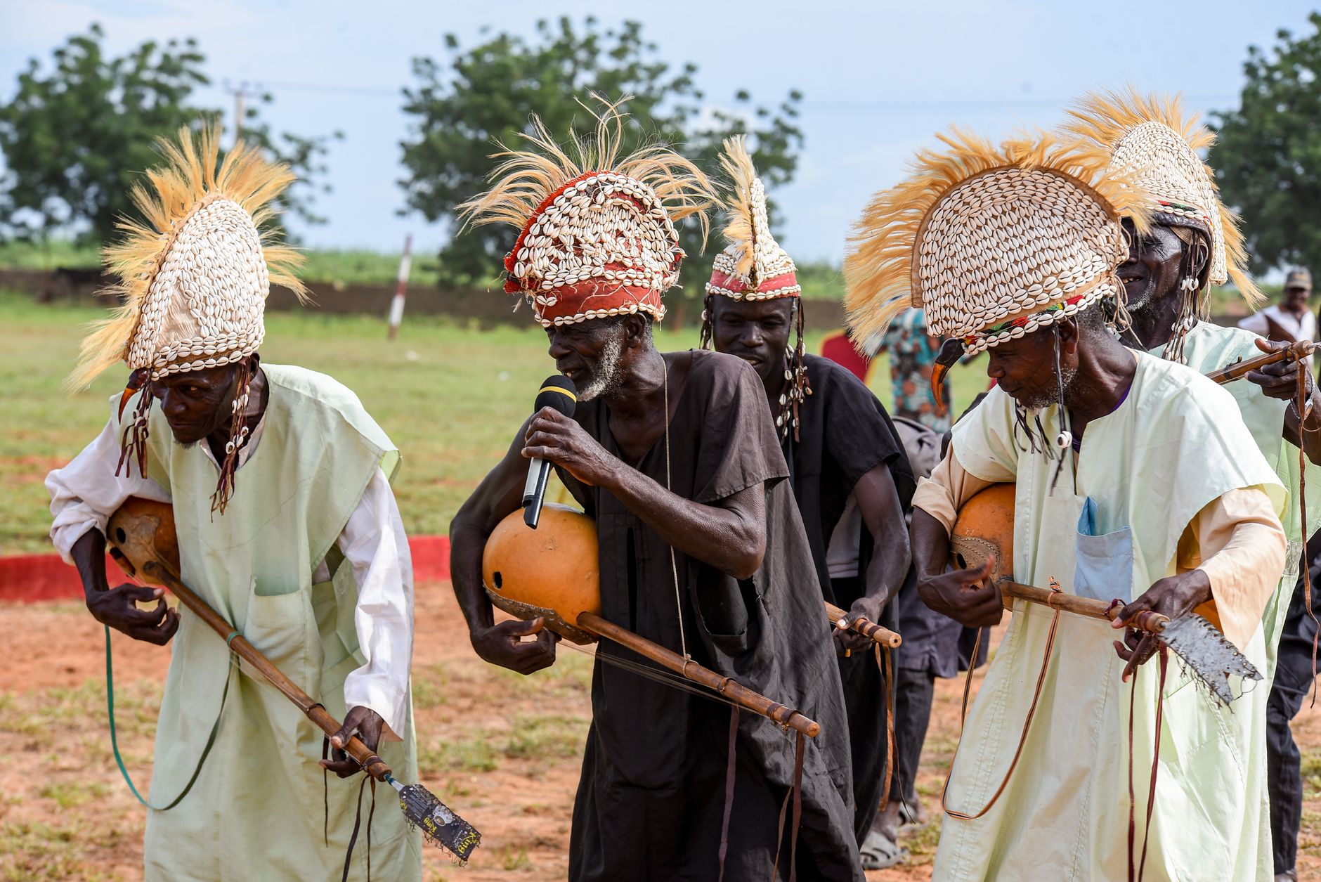 African musicians dressed in traditional attire perform with handmade instruments outdoors.