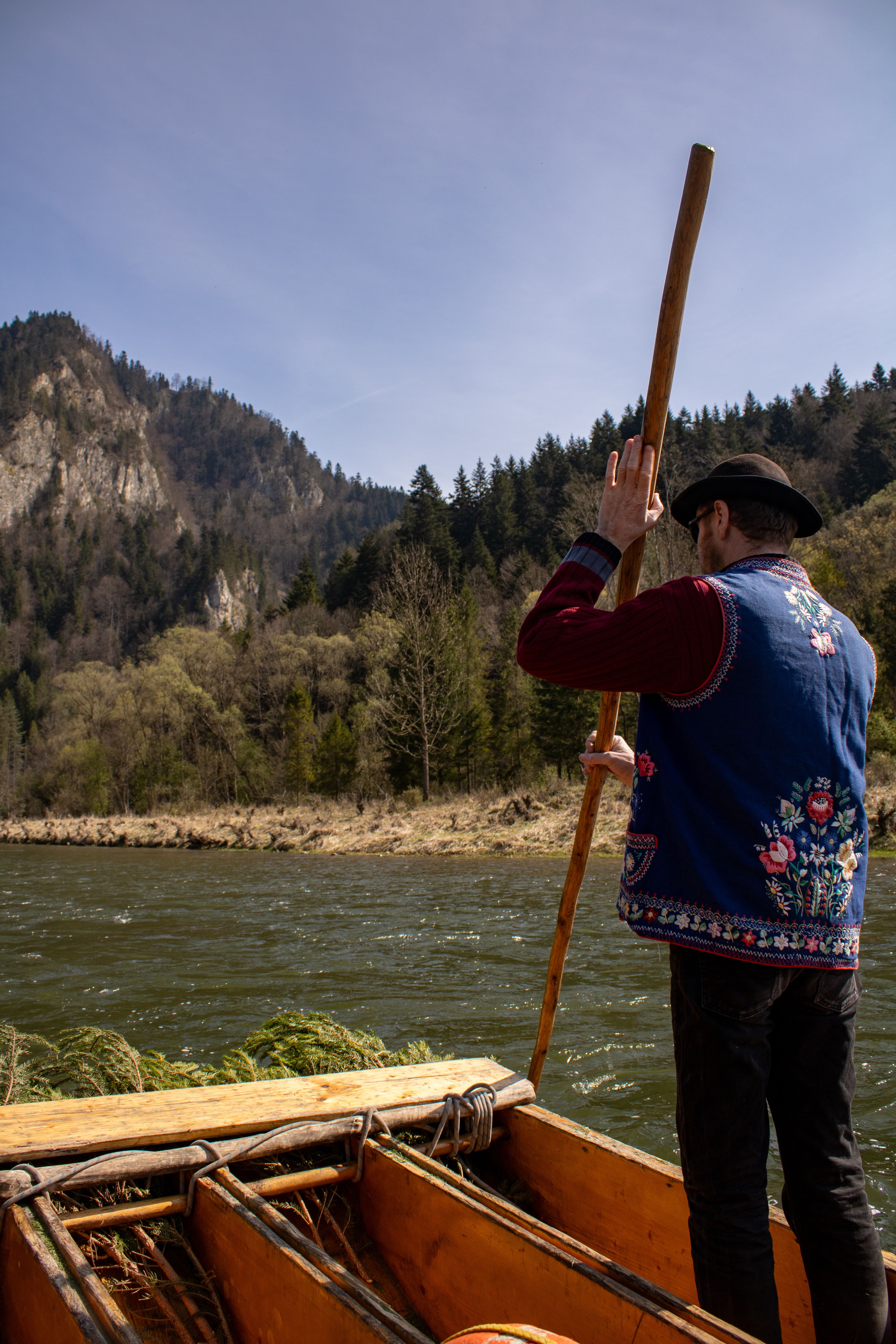 A boatman in traditional attire guides a wooden raft on a river surrounded by lush scenic mountains.