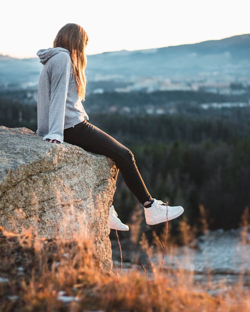 Женщина в сером худи и черных штанах сидит на скале