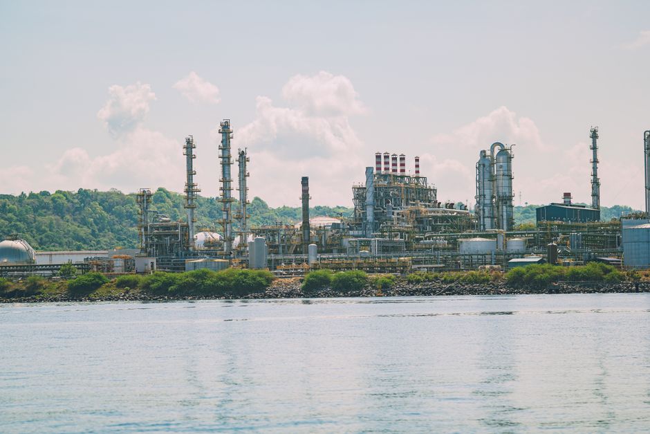 A large oil refinery situated by the water with bright green foliage in the background; clear blue skies and clouds overhead.