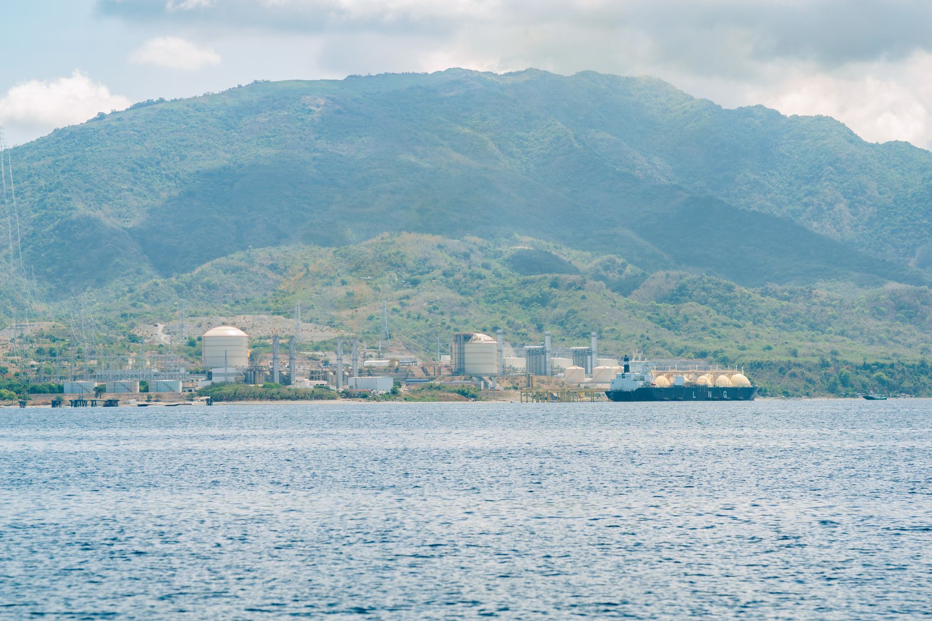 Illustrative photograph: Coastal industrial facility with a tanker ship against a mountainous backdrop.