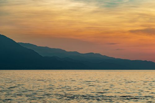 Free A peaceful sunset scene with mountain silhouettes against a colorful sky reflected on a calm sea. Stock Photo