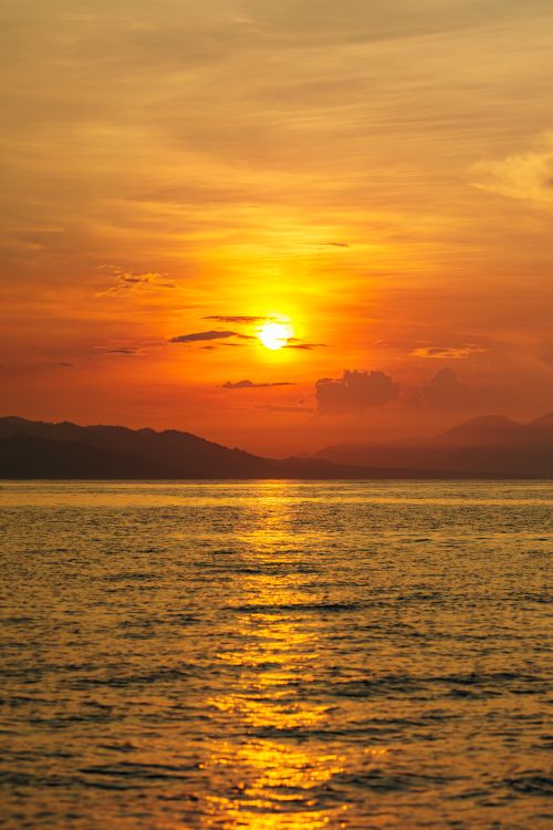 Free A stunning sunset with vibrant orange hues reflecting over the ocean water and distant mountains. Stock Photo