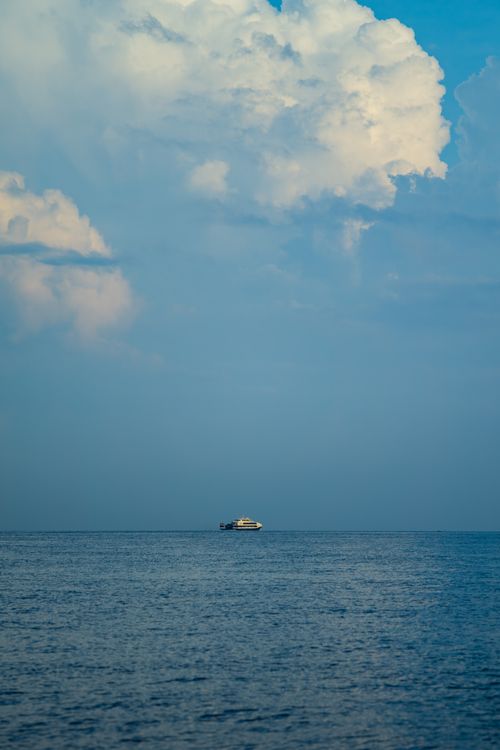 Free A lone boat floats peacefully on a vast blue ocean under a dramatic cloudy sky. Stock Photo