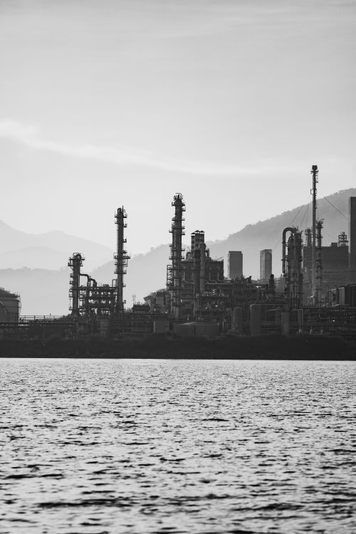 Free Black and white image of a refinery by the sea, with mountains in the background. Stock Photo
