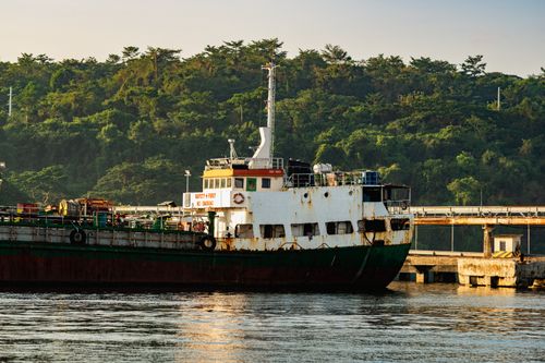 Free A weathered cargo ship docked at an industrial port, surrounded by lush green trees under clear skies. Stock Photo