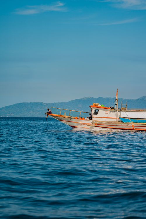 Free A fishing boat sails on calm ocean waters under a clear blue sky, with distant mountains in the background. Stock Photo