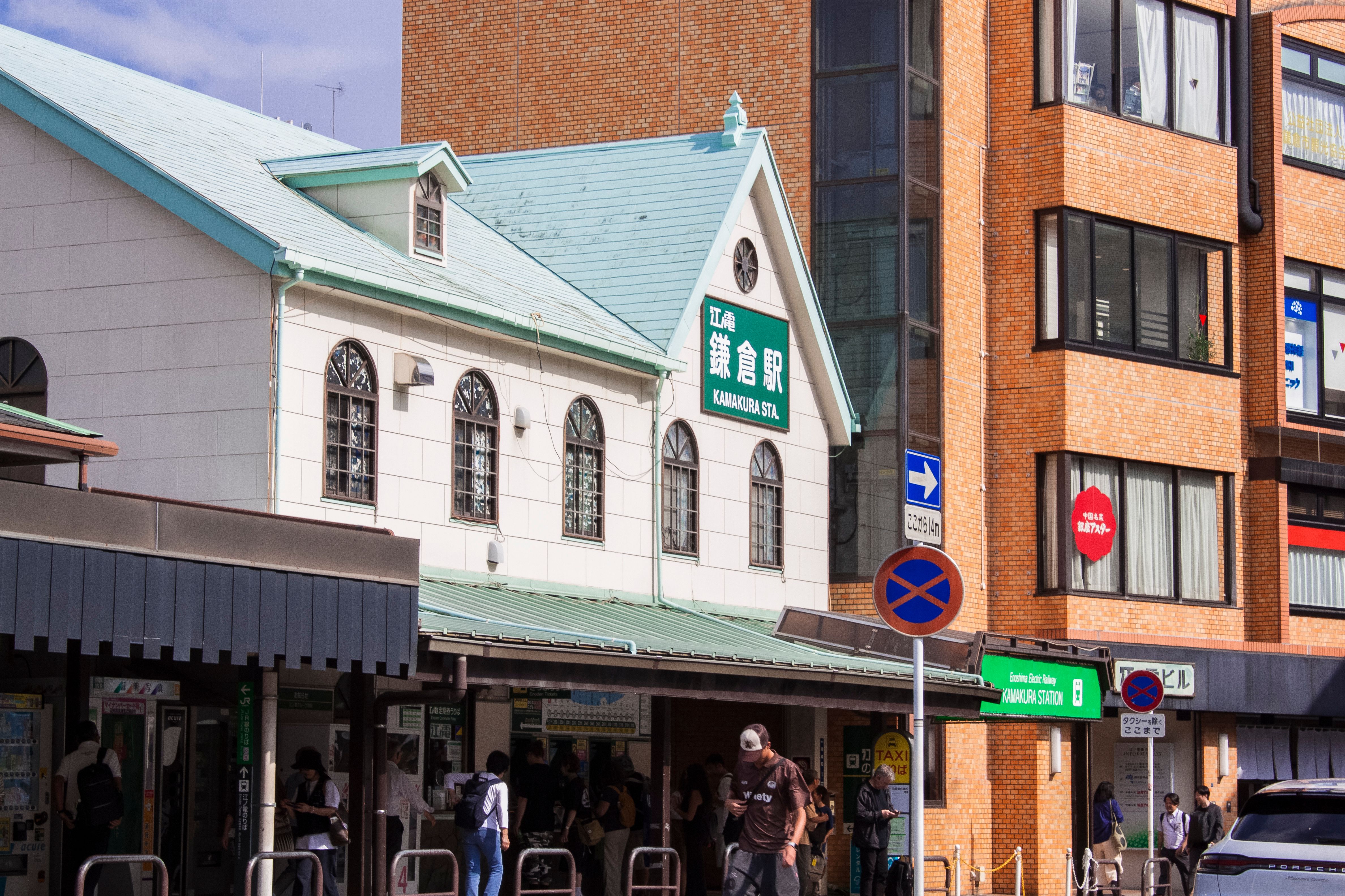 A busy entrance to Kamakura Station on a sunny day, capturing commuters and the station's architecture.