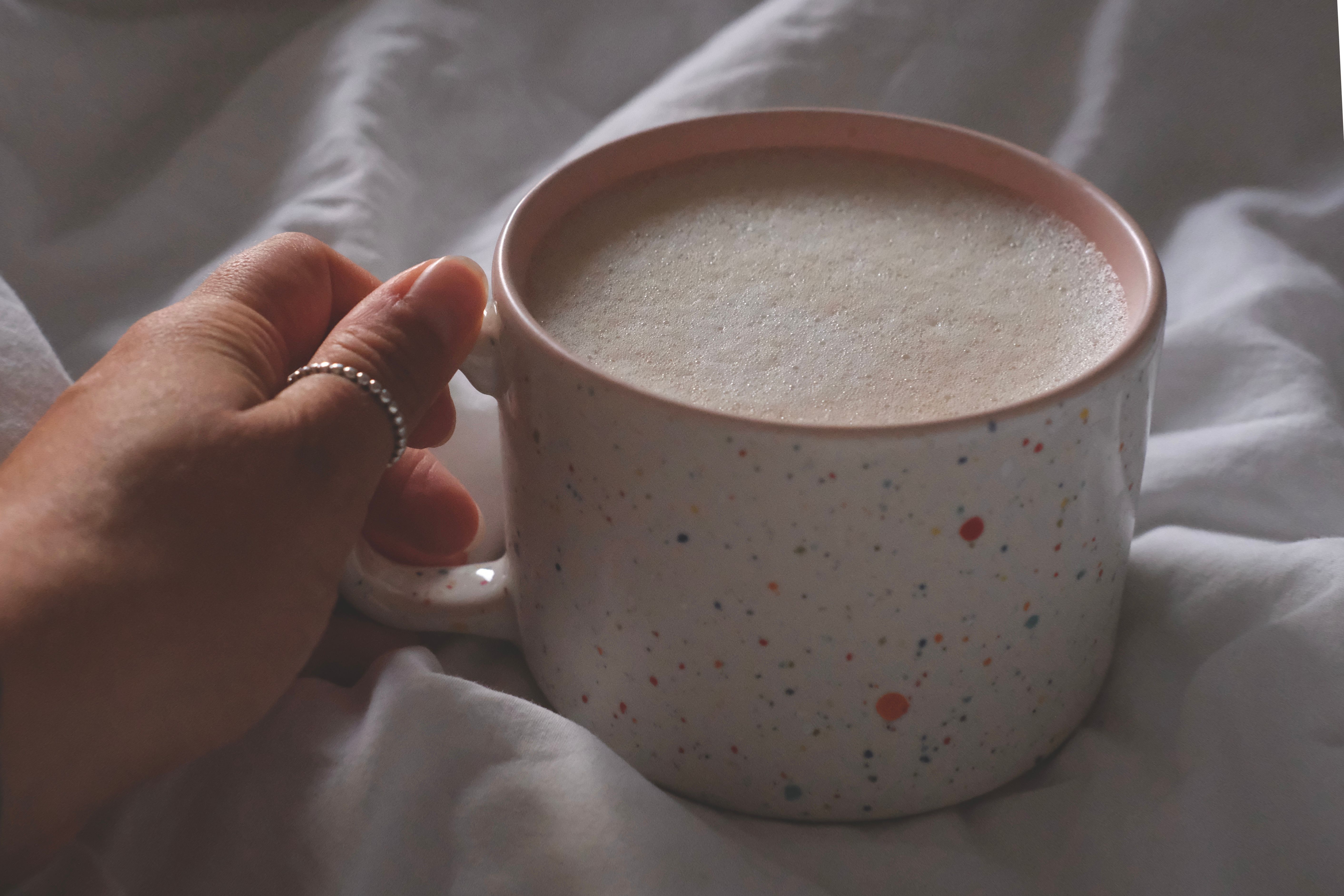 Hand holding a speckled mug filled with frothy coffee, cozy indoor setting.