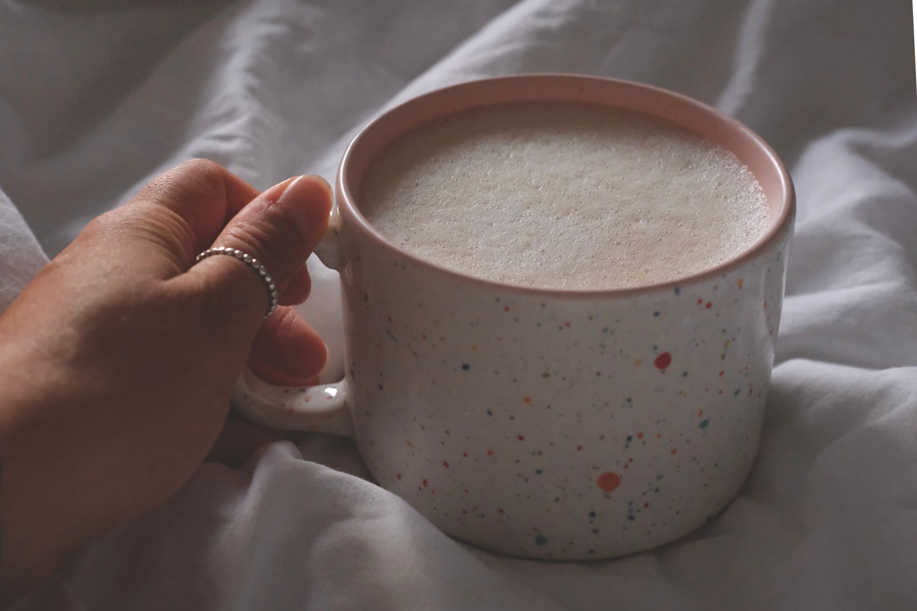 Hand holding a speckled mug filled with frothy coffee, cozy indoor setting.
