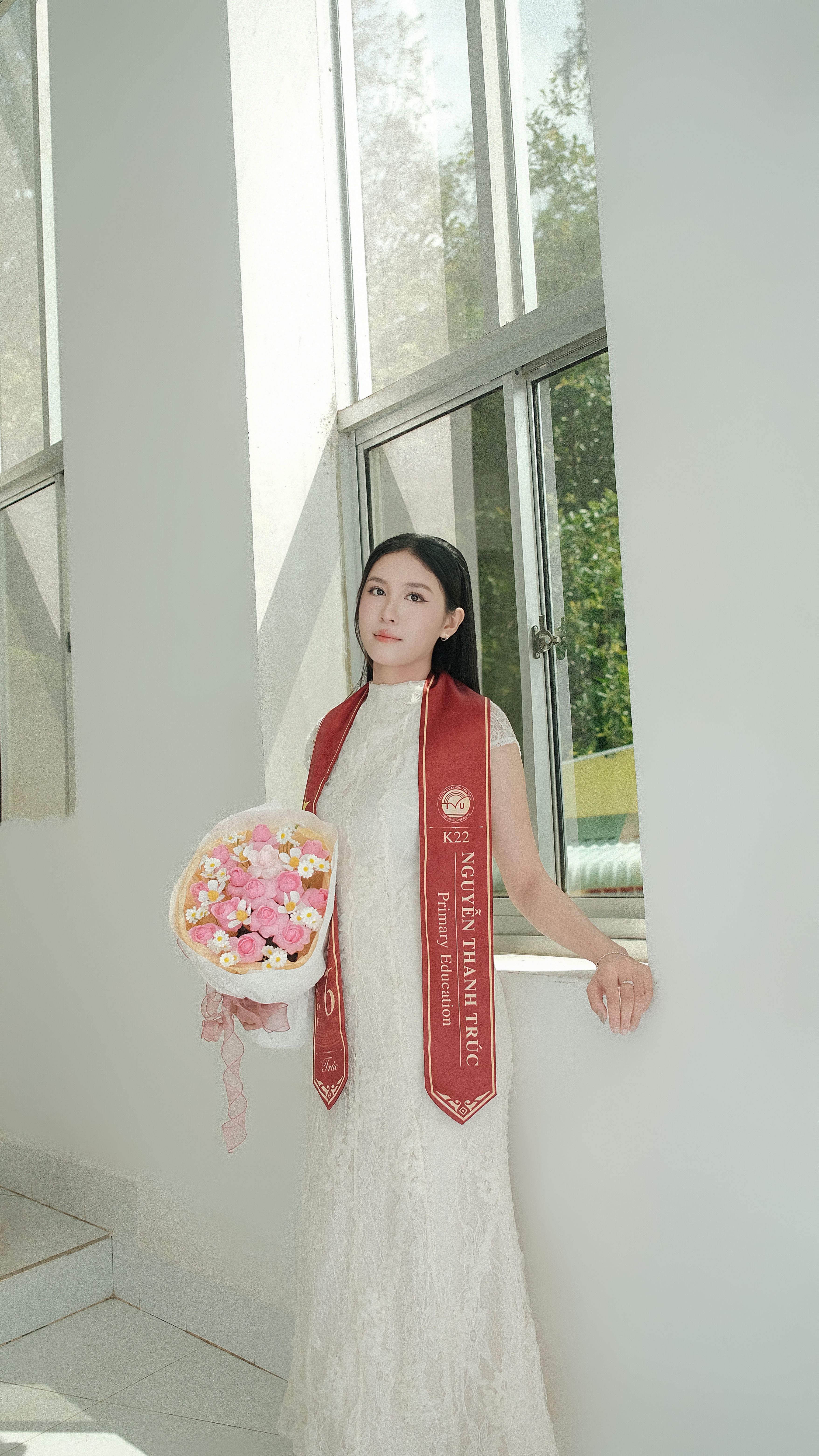 A young graduate in a white dress poses with a bouquet of flowers, standing beside a window in a sunlit hallway.