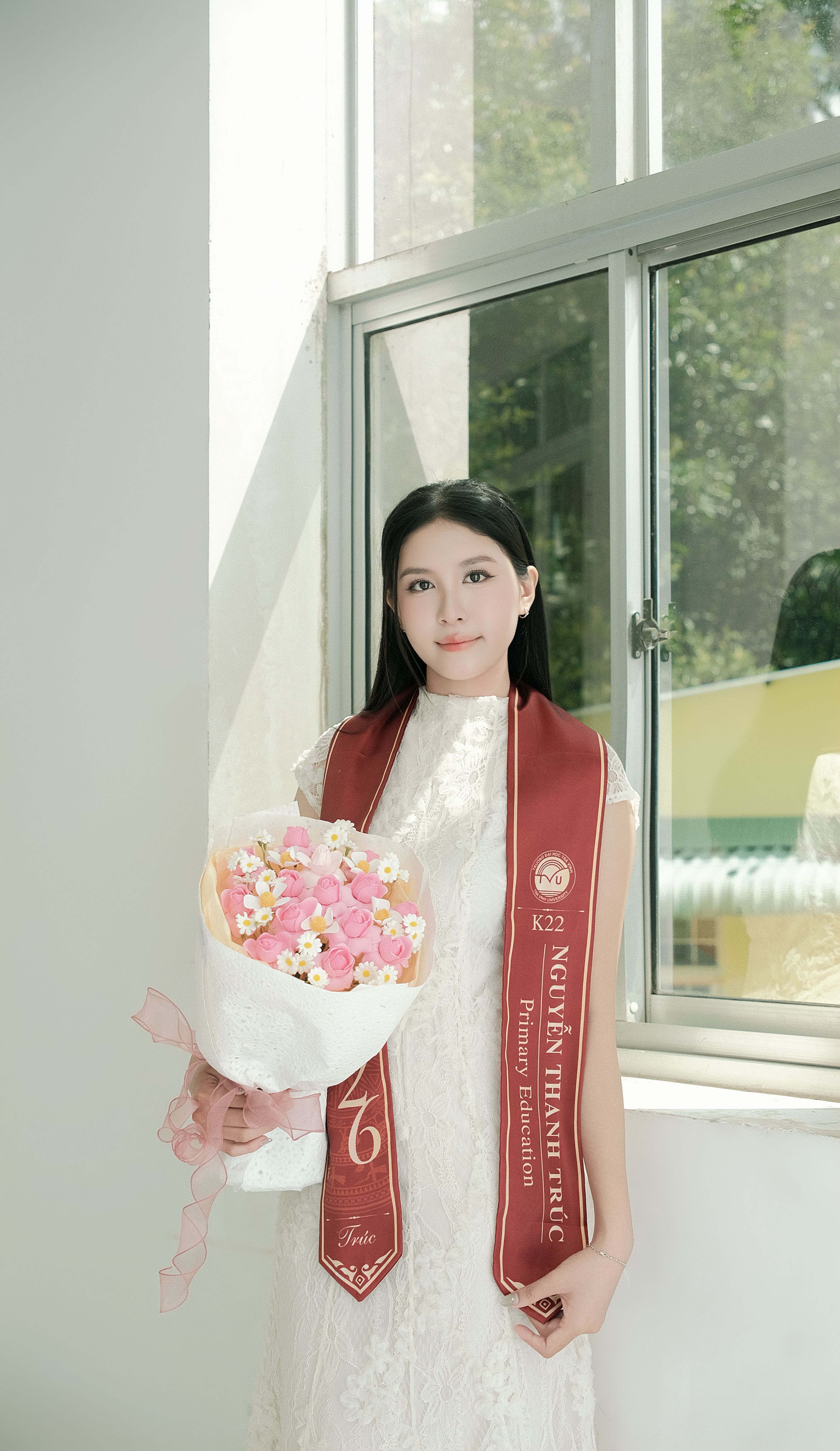 Young woman in a white dress holding flowers, wearing a graduation sash indoors.