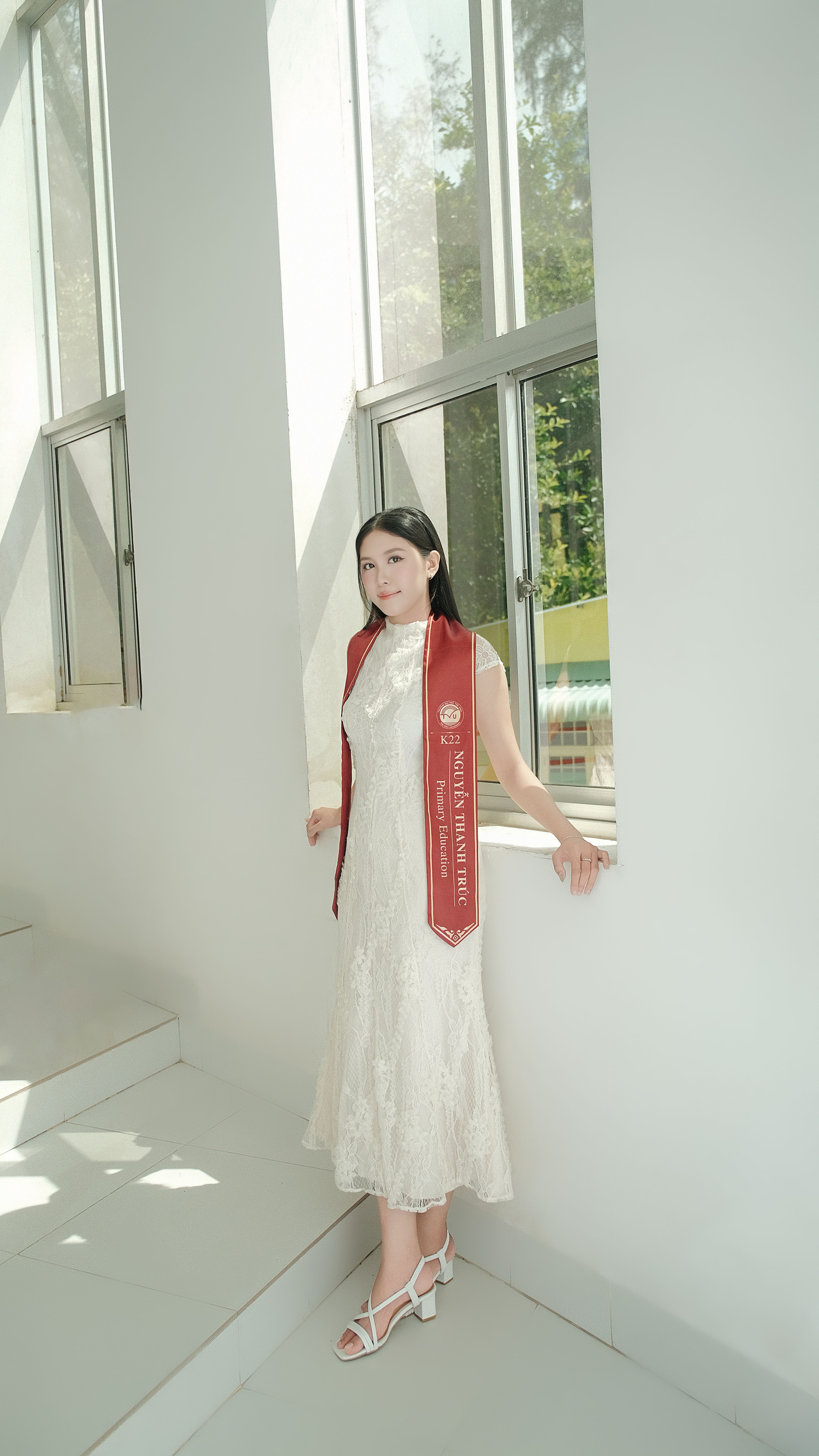 A young graduate in white gown and red sash standing by a bright window, celebrating academic success.