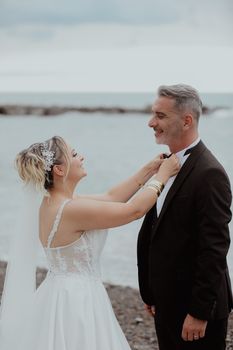 A joyful bride adjusts her groom's bow tie by the sea on a cloudy day.