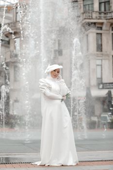 A bride in a white gown poses gracefully by a city fountain.