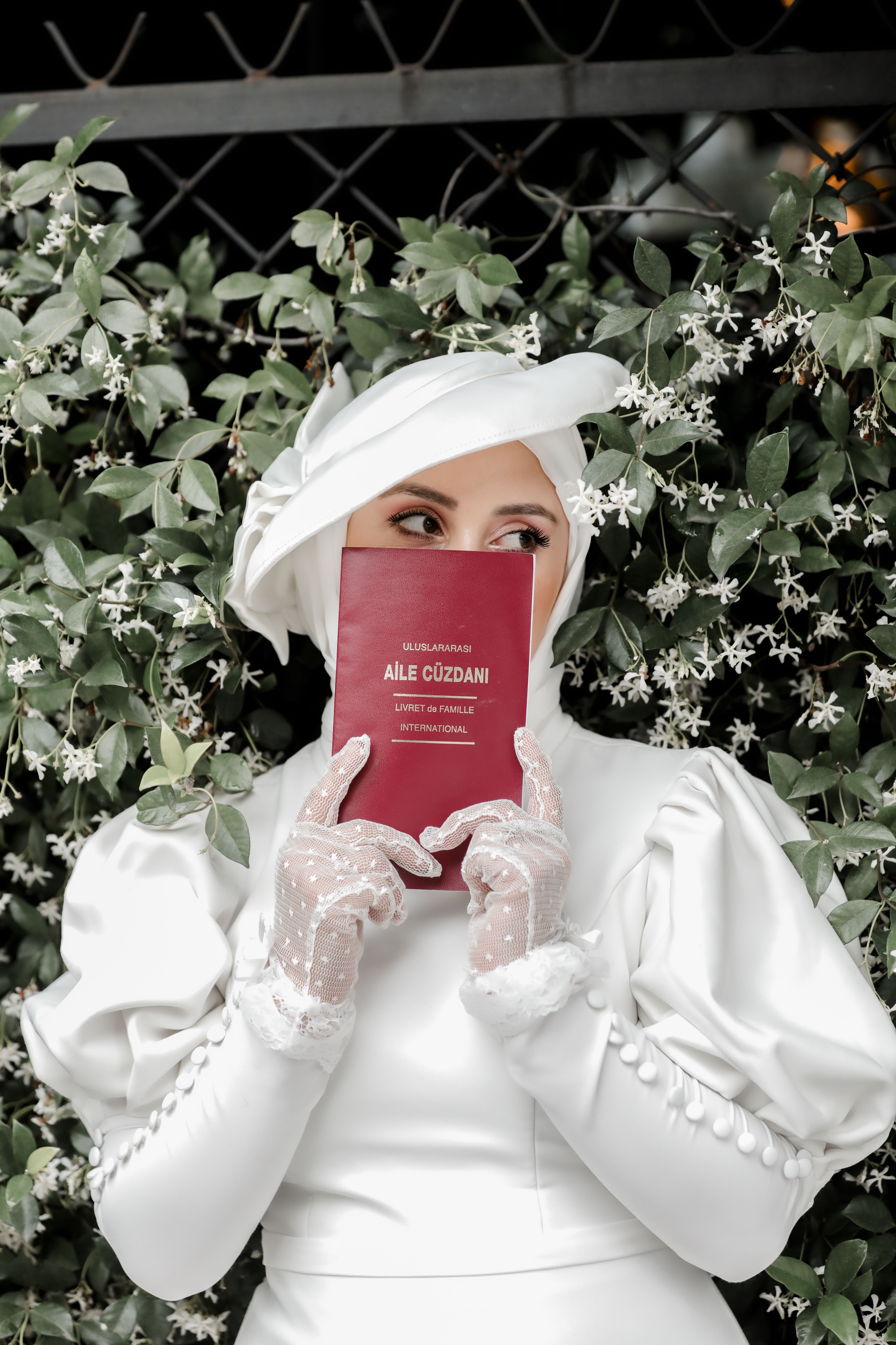A bride in a white dress holds a marriage book, surrounded by blooming flowers in an outdoor garden setting.