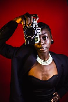 Stylish woman in black attire holding a vintage camera against a vibrant red backdrop.