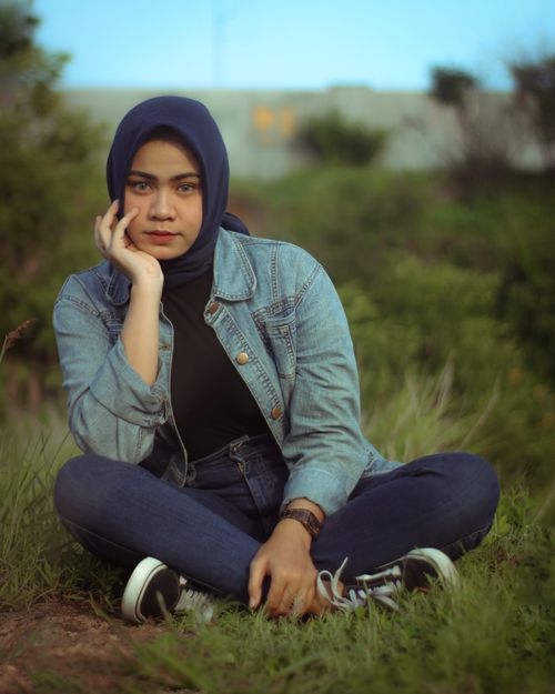 Free Serious young ethnic female in babushka sitting on grass under blue sky in summer day and looking at camera pensively Stock Photo