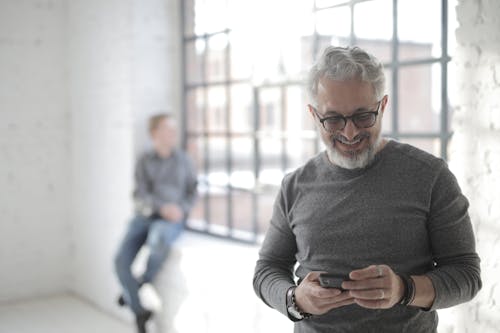 Free Middle aged bearded man in casual clothes standing with mobile phone while looking at screen and smiling during break in modern workspace Stock Photo