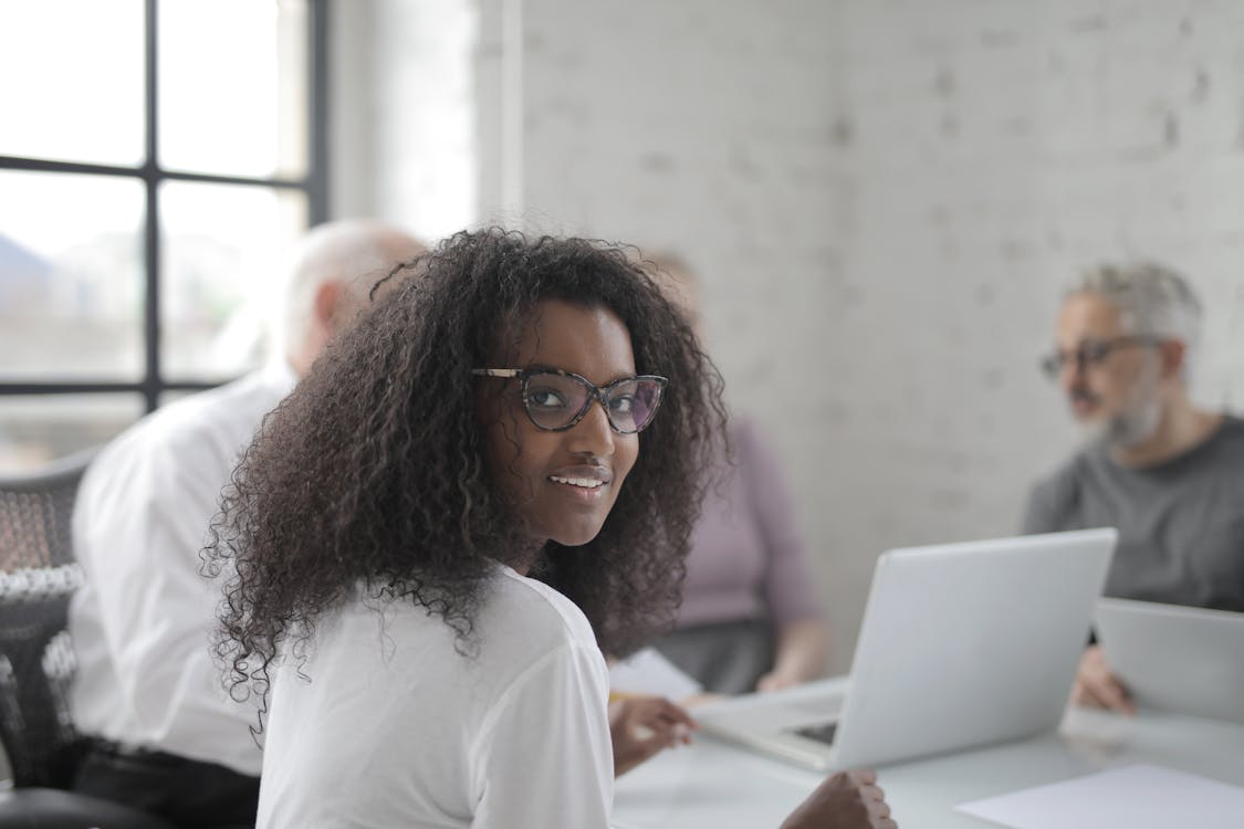 Free Positive ethnic woman wearing formal clothes and eyeglasses smiling while working with colleagues in modern workspace and looking at camera Stock Photo