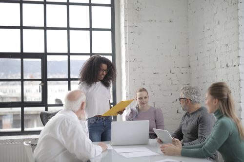 Free Concentrated ethnic woman reading documents while standing near table and explaining details of project to colleagues in modern office near window Stock Photo