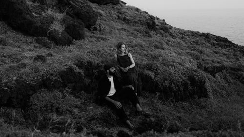 Free Black and white photo of a couple sitting on a rocky cliff overlooking the ocean. Stock Photo