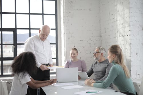 Serious elderly man wearing formal clothes standing while explaining business project with colleagues and discussing details during brainstorm at modern office