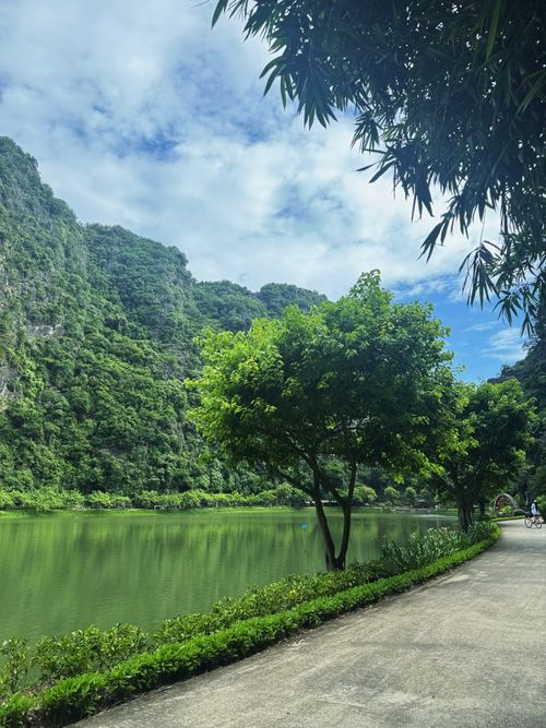 Free A serene riverside pathway winds through a lush green landscape under a bright sky. Stock Photo