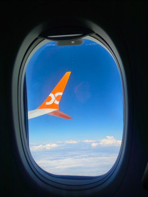 View from an airplane window showing blue sky and clouds during mid-flight with visible wing.