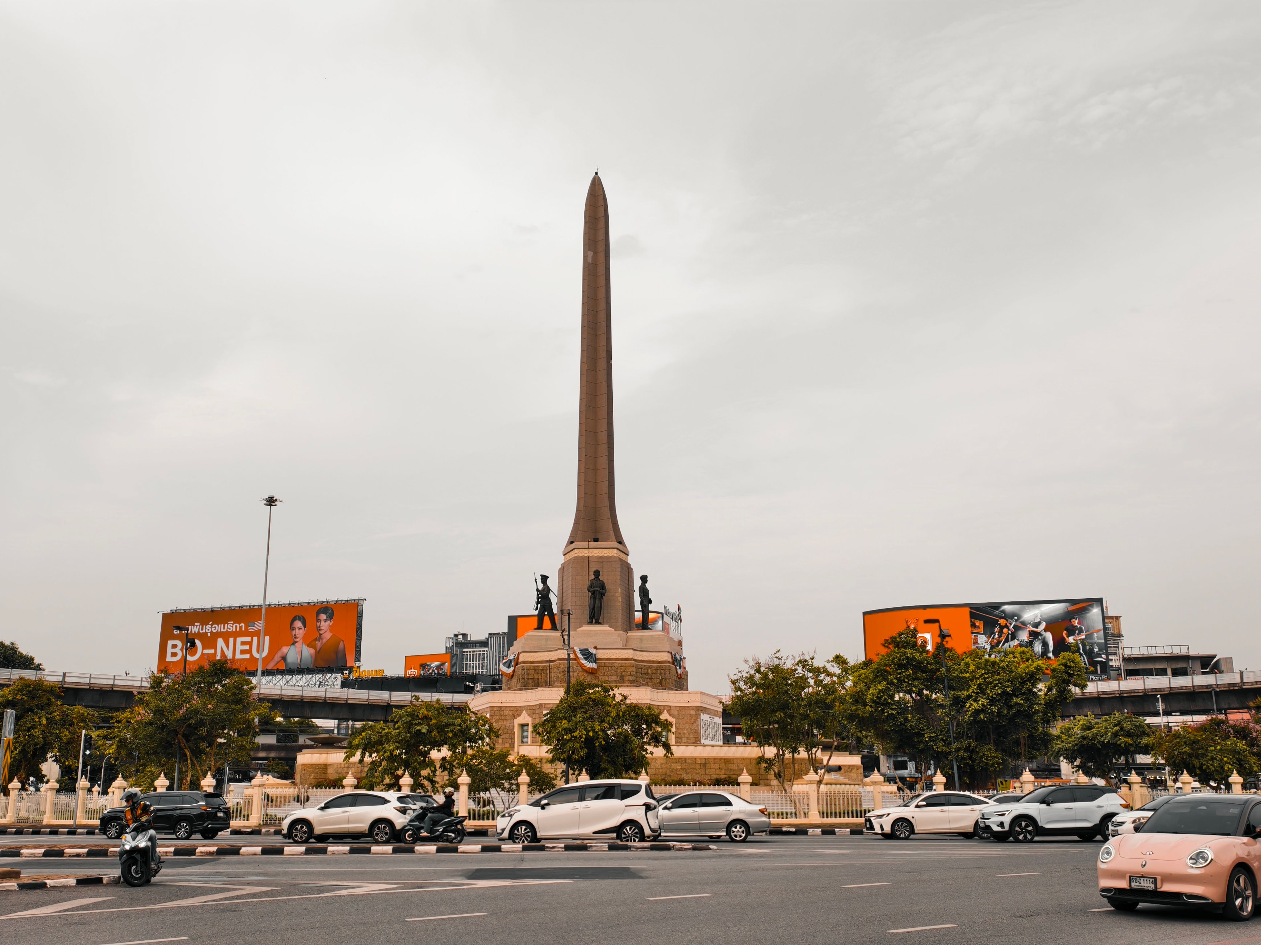 The iconic Victory Monument in Bangkok, Thailand, surrounded by city traffic and billboards.